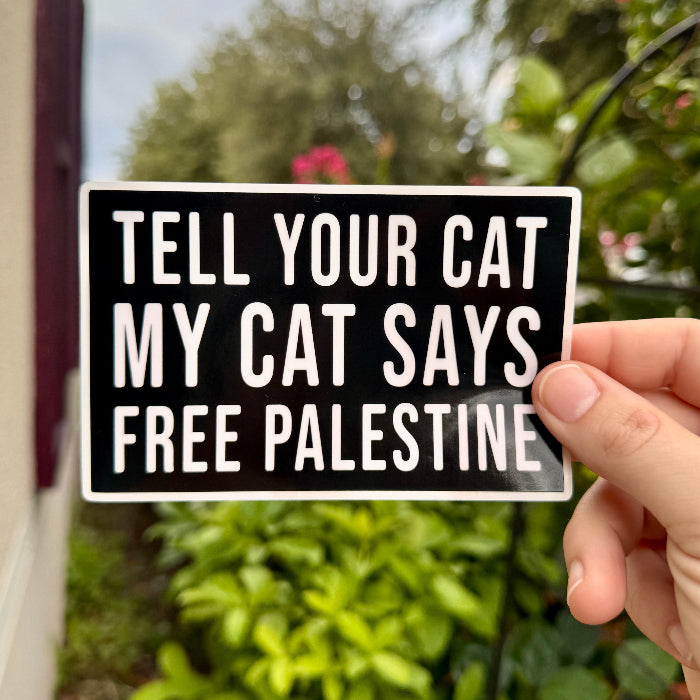 Hand holding a sign with text 'Tell your cat my cat says free Palestine' against a blurred green outdoor background.